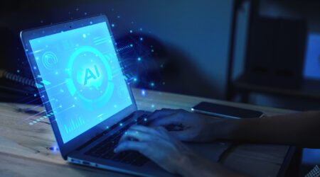 A person typing on a laptop with a glowing screen displaying "AI" and digital graphics in a dimly lit environment.