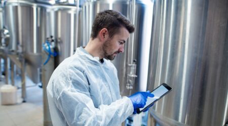 A person in a lab coat and gloves is using a tablet near metal fermentation tanks in a brewery or laboratory setting.
