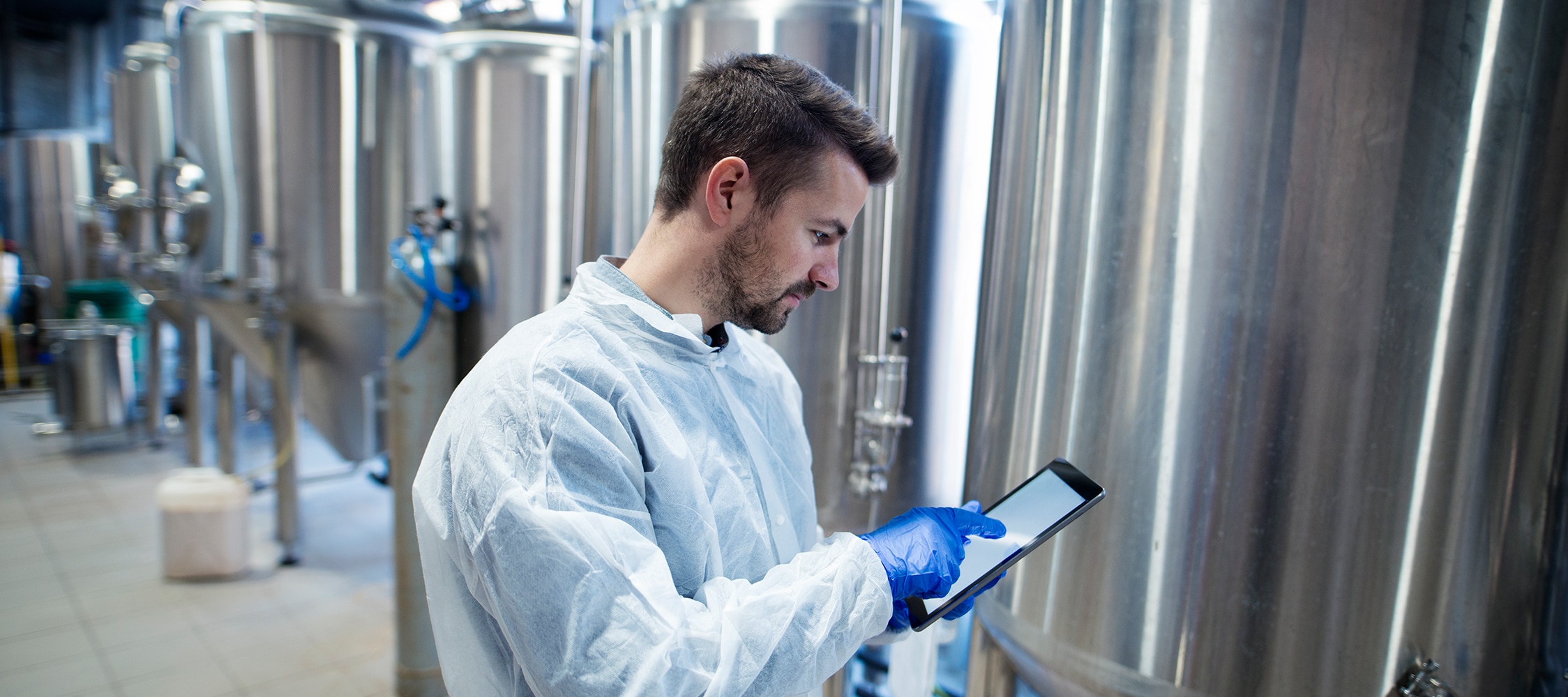 A person in a lab coat and gloves is using a tablet near metal fermentation tanks in a brewery or laboratory setting.