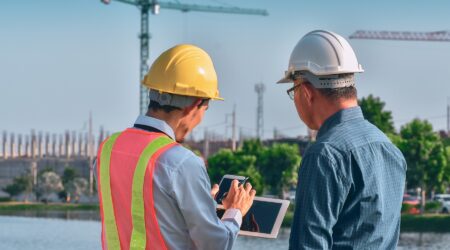 Two construction workers, one in a yellow hard hat and safety vest, are discussing plans on a tablet at a construction site.