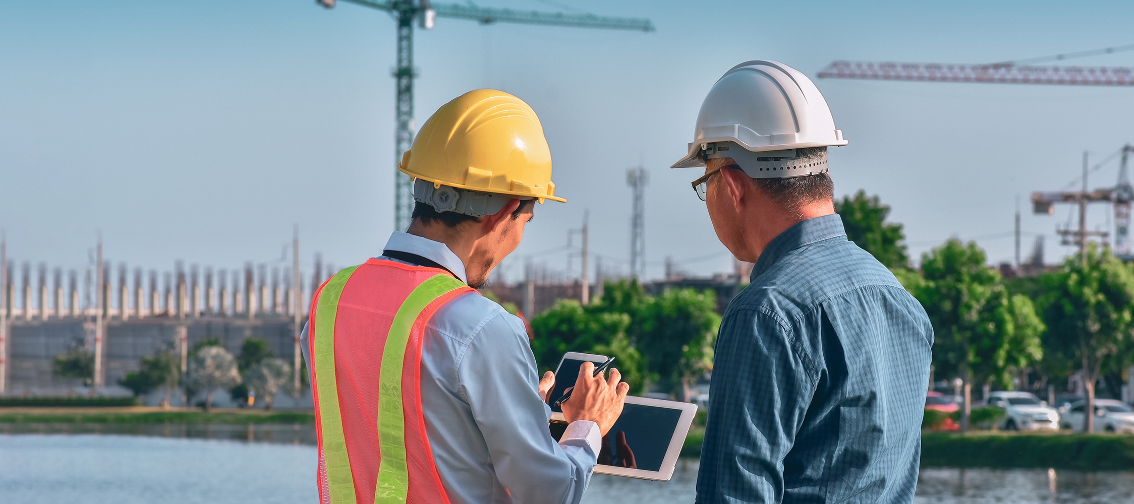 Two construction workers, one in a yellow hard hat and safety vest, are discussing plans on a tablet at a construction site.