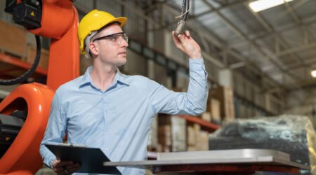 A man in a hard hat operates a robotic arm in an industrial setting. He holds a tool while inspecting the robot's movement.