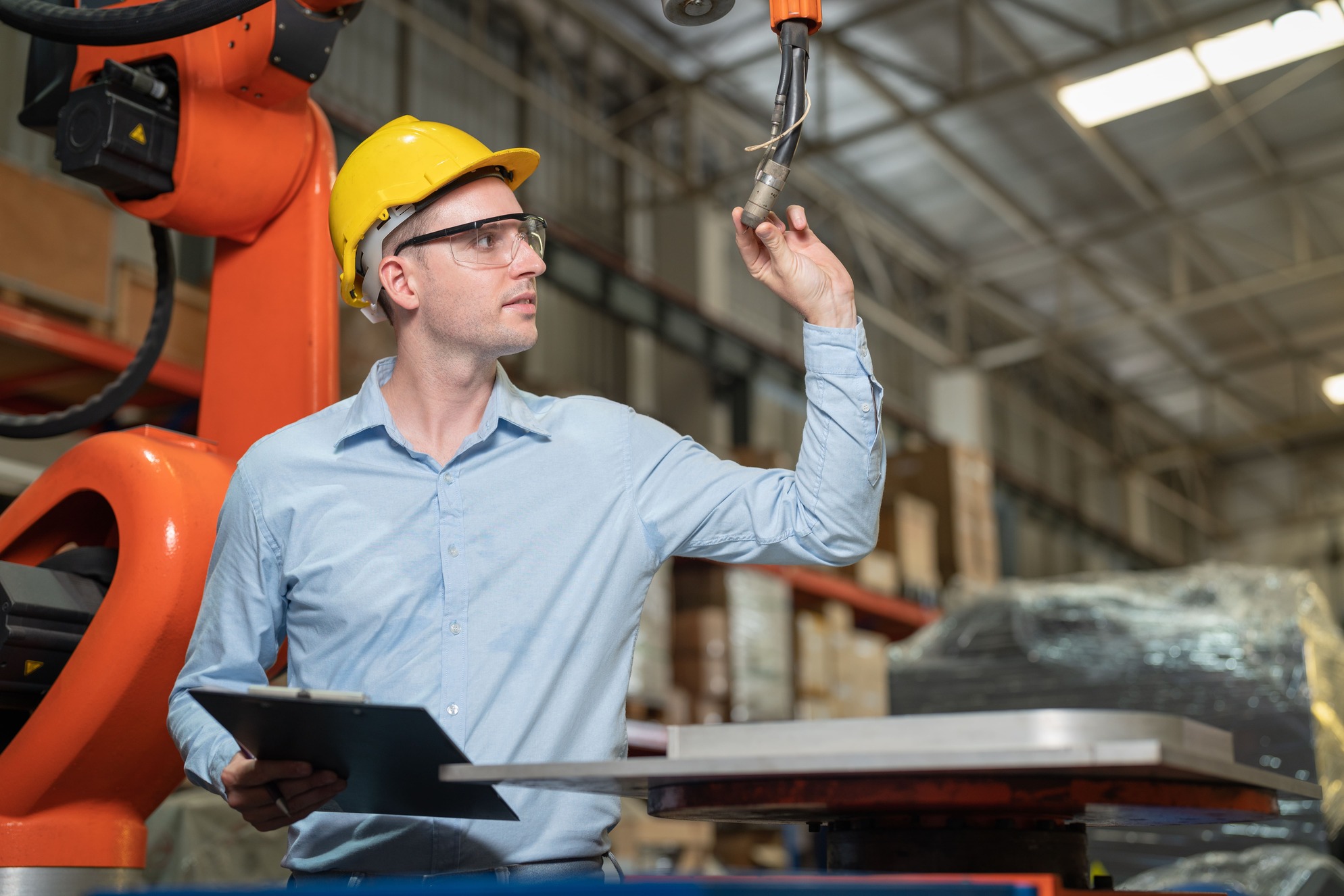 A man in a hard hat operates a robotic arm in an industrial setting. He holds a tool while inspecting the robot's movement.