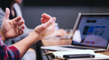 A person gesturing with their hands at a table with a laptop and notebook.