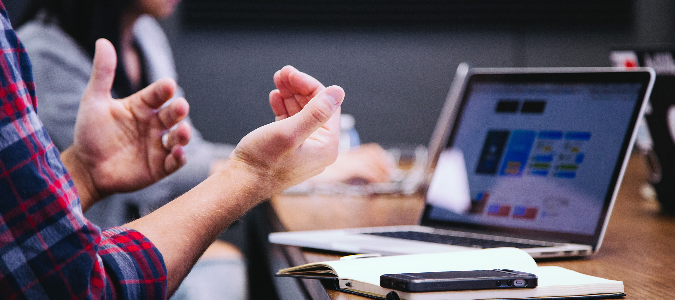 A person gesturing with their hands at a table with a laptop and notebook.