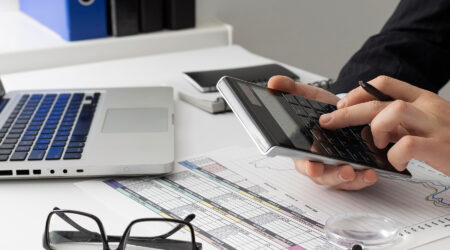 Person using a calculator with a laptop and glasses on the desk.