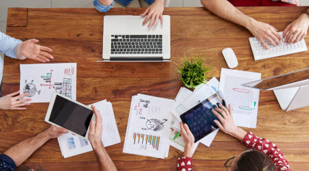 People around table in office with laptops, tablets, and data projections.