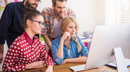 Group of coworkers gathered around a computer, collaborating for some task in a bright modern office.