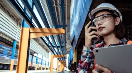 A woman in a safety helmet and glasses uses a walkie-talkie while holding a tablet, standing in a warehouse or industrial setting.