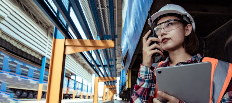 Woman in safety helmet and glasses using walkie-talkie and holding tablet in industrial setting.
