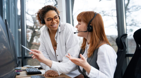 Two women on a customer Support team wearing headsets and talking to each other while looking at a desktop computer.