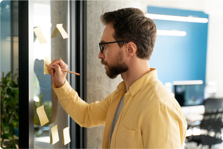 A man is writing on a sticky note and placing it on a glass wall in an office setting. He is wearing glasses and a yellow shirt.