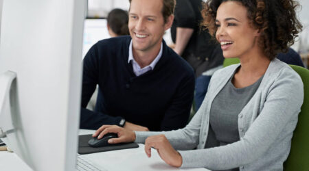 Man and woman sitting next to each other in an office looking at computer monitor and smiling.