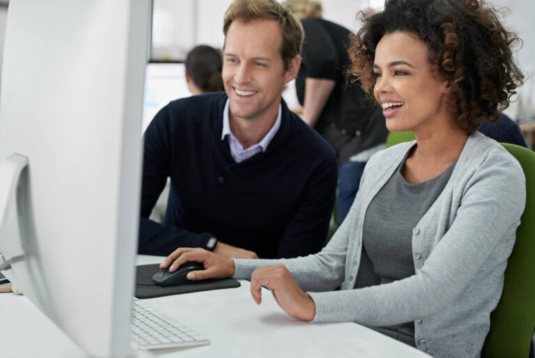 Man and woman sitting next to each other in an office looking at computer monitor and smiling.
