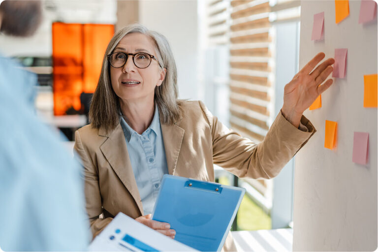A woman with gray hair and glasses is discussing with a person off-camera, gesturing towards a wall with colorful sticky notes. She is holding a clipboard.