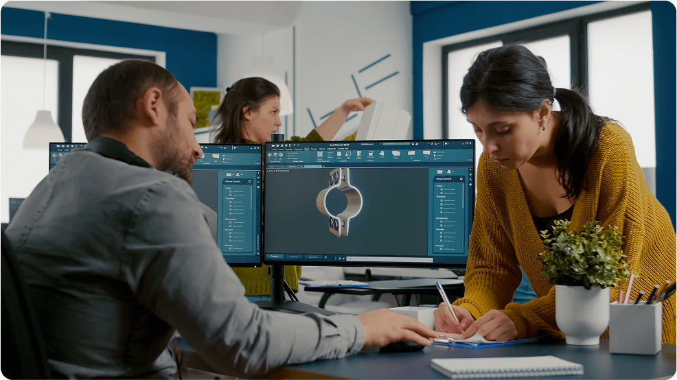 A man and a woman are collaborating in an office. The man, seated at a desk with two computer monitors displaying a design, is writing notes. The woman, standing, is holding a document and assisting him. The workspace has a modern design with light colors.