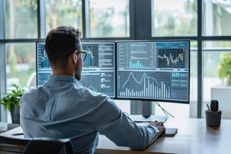 Person at desk using multiple monitors showing data and graphs, with plant and smartphone on desk.