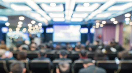 A blurred image of an audience seated in a conference room, facing a screen.