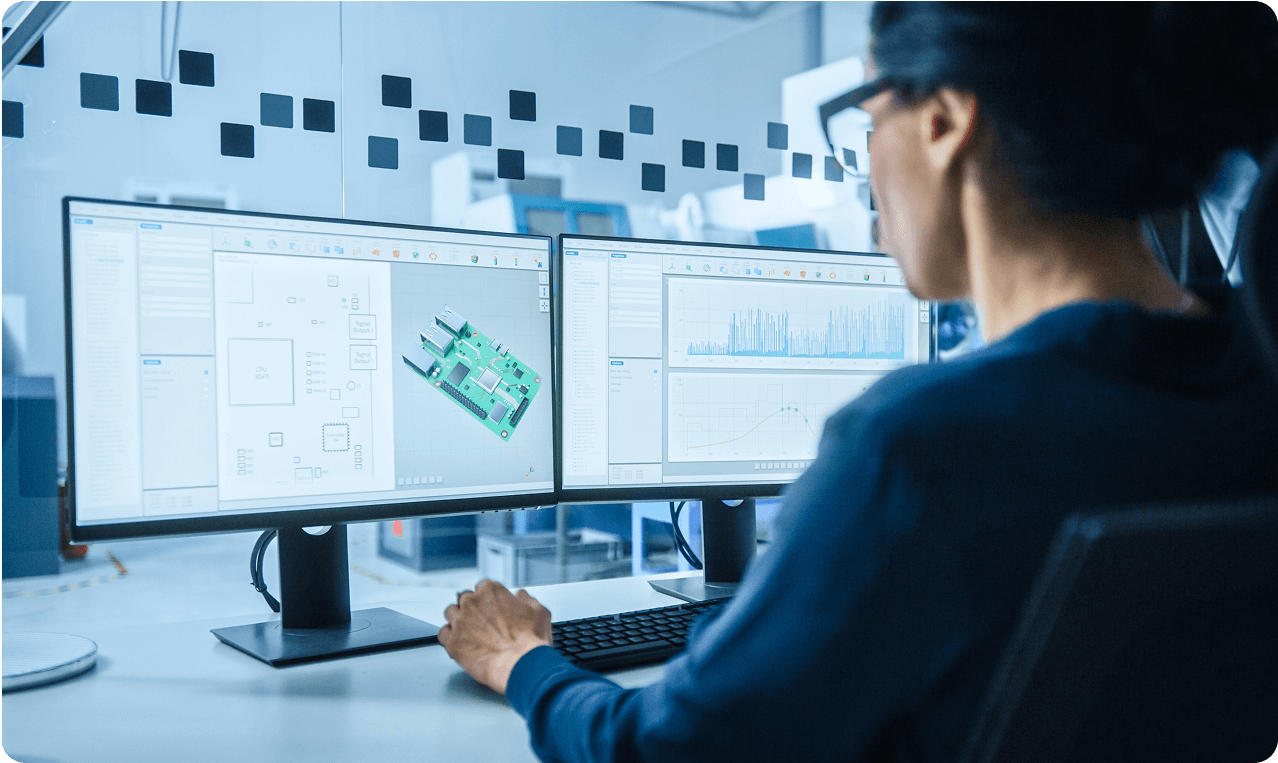 A person is seated at a desk using two computer monitors displaying a circuit board design and data graphs.