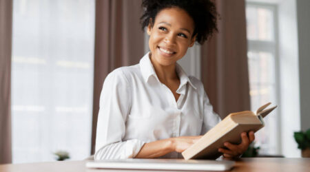 Woman sitting at a desk and holding a book while smiling looking into distance.