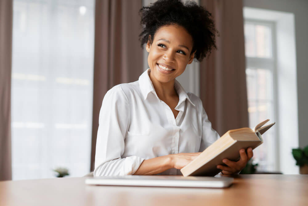 Woman sitting at a desk and holding a book while smiling looking into distance.