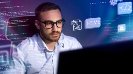 A man wearing glasses sits at a desk, looking at a computer screen with HTML code and graphics related to web development displayed around him.
