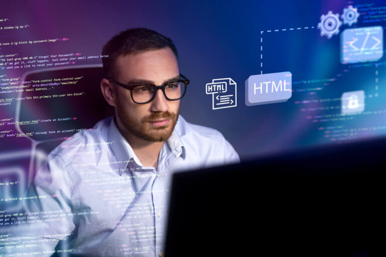 A man wearing glasses sits at a desk, looking at a computer screen with HTML code and graphics related to web development displayed around him.