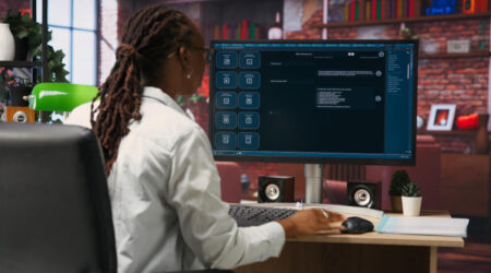 A person in a white shirt sits in front of a computer monitor displaying an AI conversation. The workspace includes a small plant and speakers, bookshelves, and window light in the background.