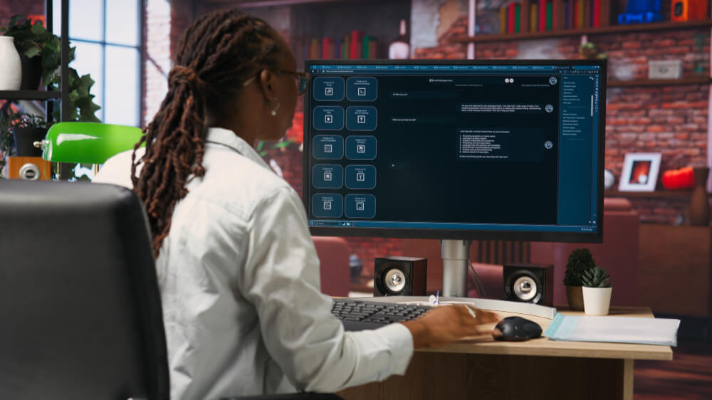 A person in a white shirt sits in front of a computer monitor displaying an AI conversation. The workspace includes a small plant and speakers, bookshelves, and window light in the background.