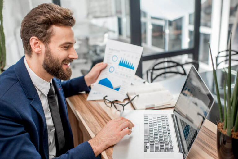 A man in a suit at a desk, smiling while looking at a laptop and holding a paper with charts and metrics on it.