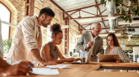 Colleagues standing around a table in an industrial workspace and looking at each others' laptops.