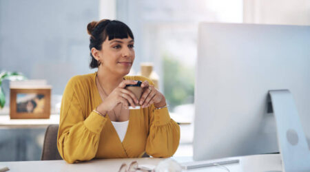 A woman holding a coffee cup while sitting at a desk and looking at her computer screen.