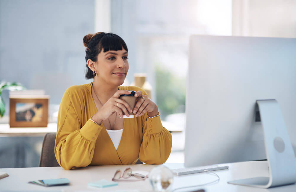 A woman holding a coffee cup while sitting at a desk and looking at her computer screen.