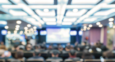 A blurred image of an audience seated in a conference room, facing a screen.