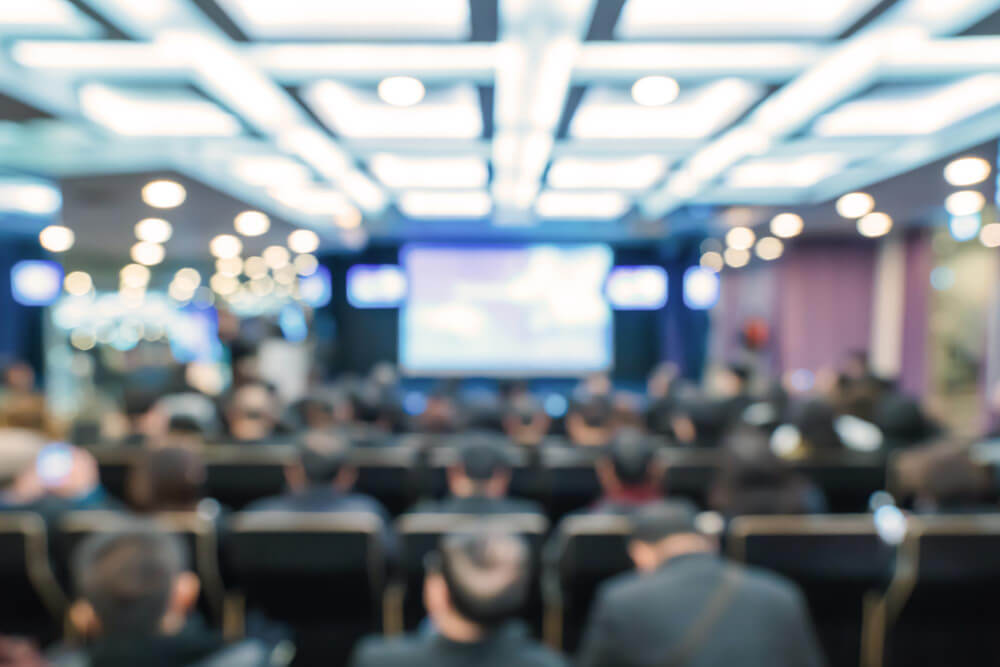 A blurred image of an audience seated in a conference room, facing a screen.