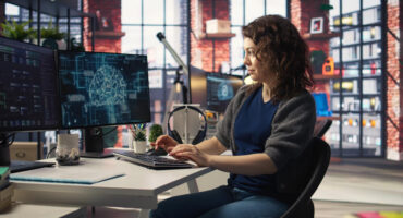 Woman in an office, typing, and looking at computer monitors that show AI.