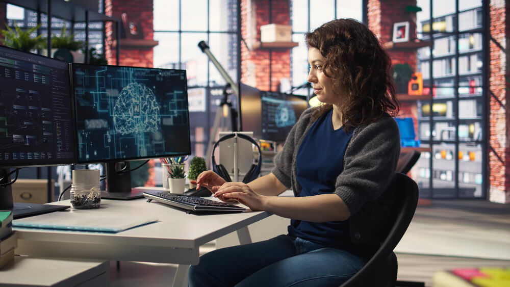 Woman in an office, typing, and looking at computer monitors that show AI.