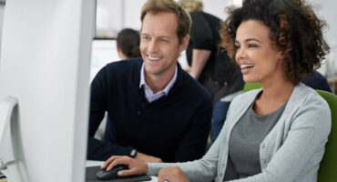 Man and woman sitting next to each other in an office looking at computer monitor and smiling.