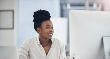 A woman with dark, braided hair sits at a desk, smiling while looking at a computer screen. A laptop, a small plant, and a cup are on the desk.