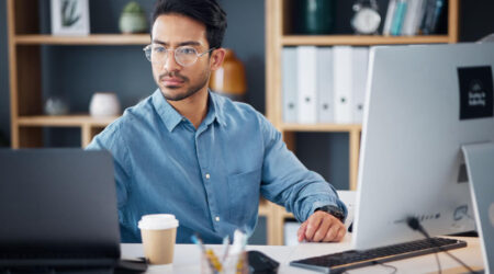 A focused individual in a blue shirt works at a desk with a laptop, desktop monitor, and a coffee cup. Bookshelves and office items are in the background.