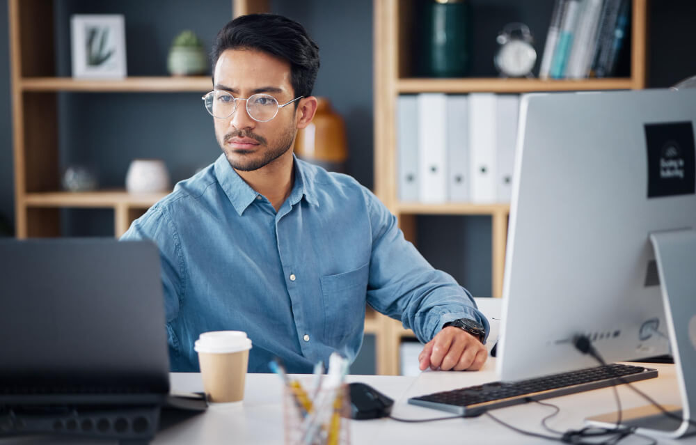 A focused individual in a blue shirt works at a desk with a laptop, desktop monitor, and a coffee cup. Bookshelves and office items are in the background.