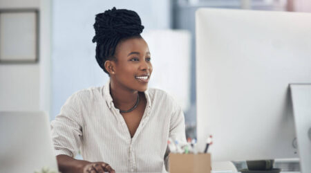 A woman with dark, braided hair sits at a desk, smiling while looking at a computer screen. A laptop, a small plant, and a cup are on the desk.