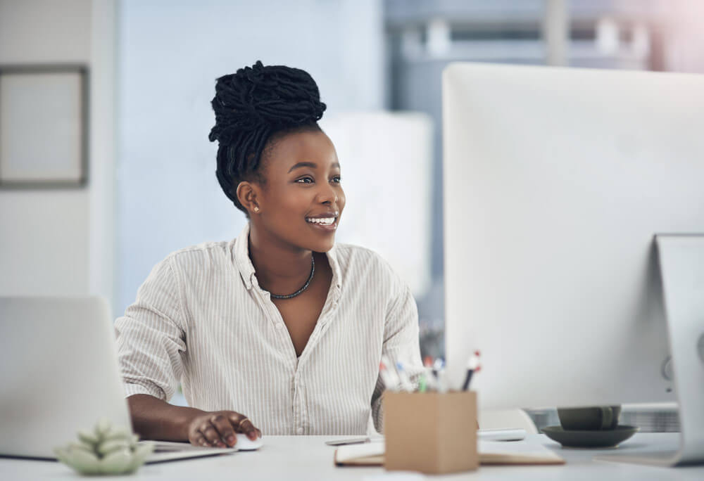A woman with dark, braided hair sits at a desk, smiling while looking at a computer screen. A laptop, a small plant, and a cup are on the desk.